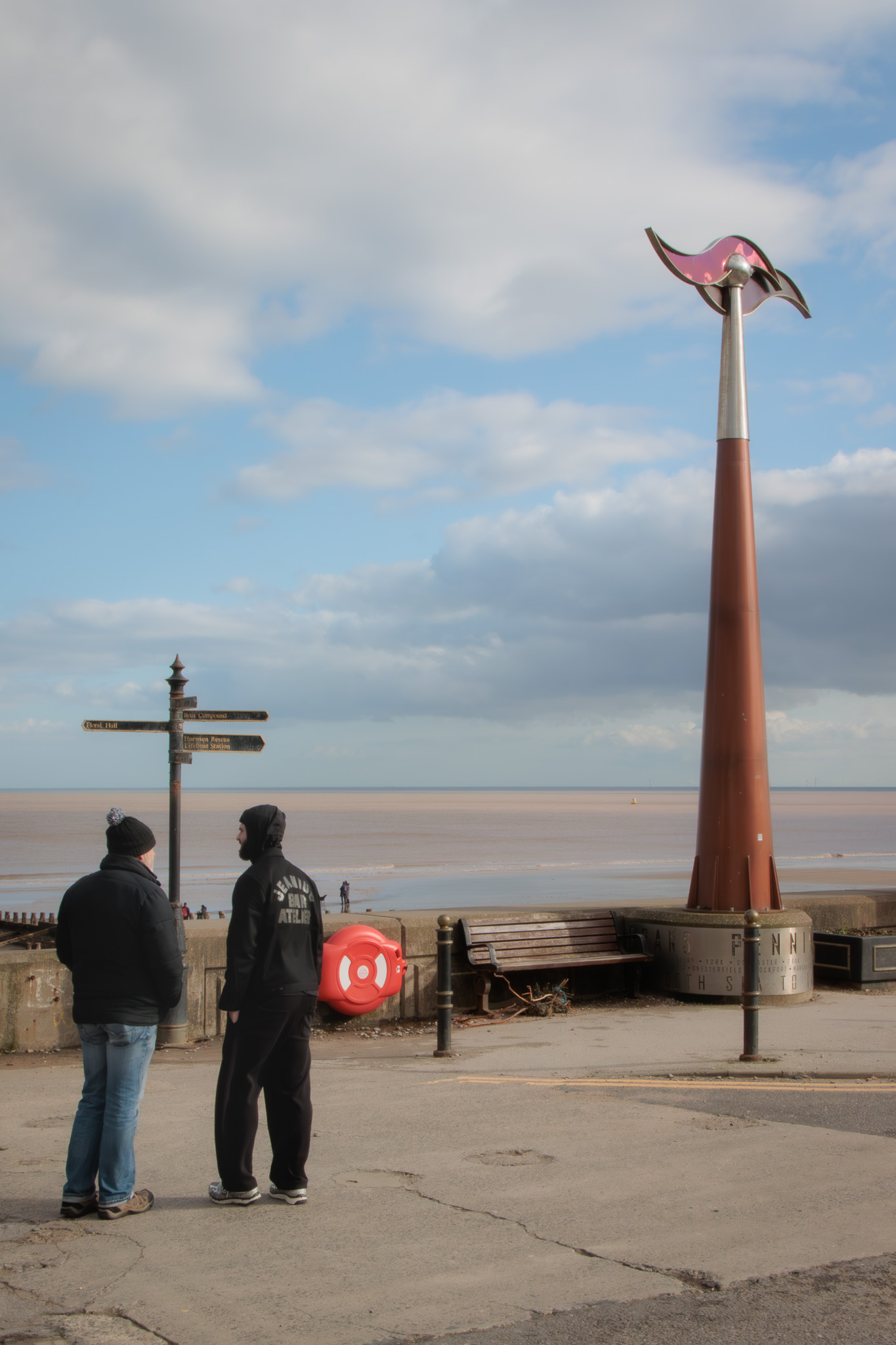 Photograph of the Hornsea seamark taken from land. The seamark is a tall tapered post, painted reddish brown and chrome, with a slupture depicting waves at the top. To the side is a signpost and in front of this two men stand with their backs to the camera. In the background is the sea, and in the far distance wind turbines can be seen.